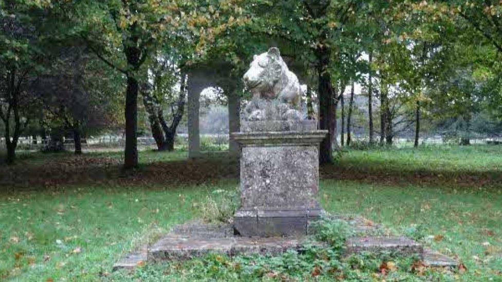 Stone statue of a lioness on a plinth, surrounded by trees, with the planned stone temple in the background