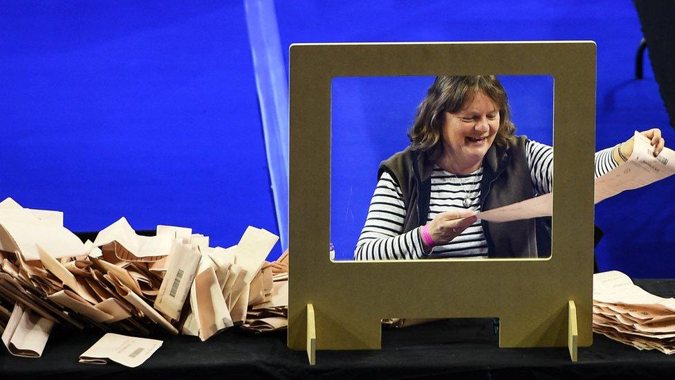 A counter smiling as she counts peach ballot papers, standing in front of a Covid shield