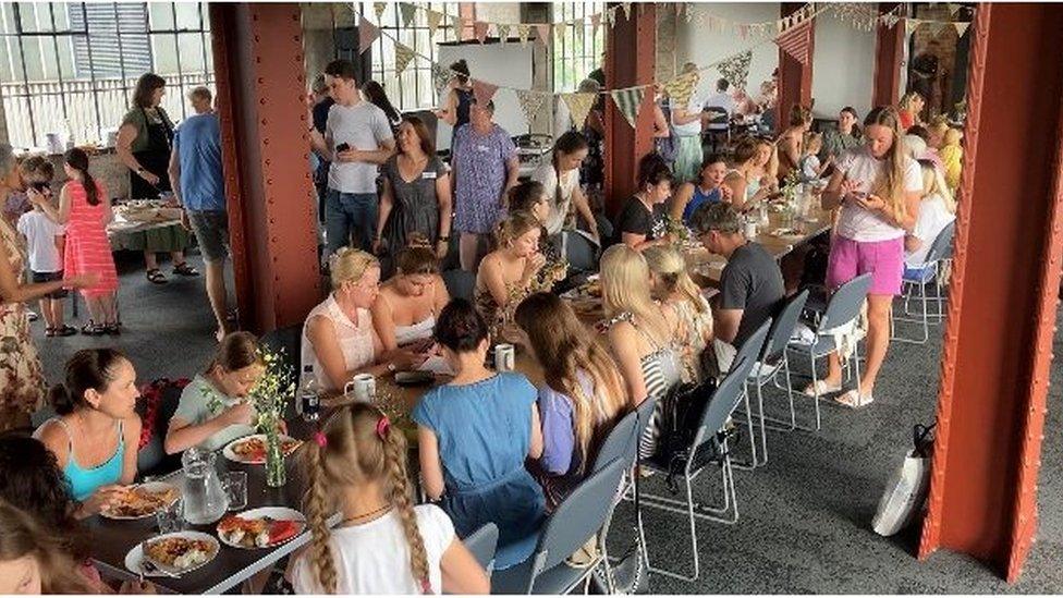 Ukrainian refugees, adults and children, eating together on a long table stretching the length of a large room at The Pattern Church in Swindon