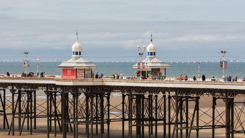 Showing two of four Victorian pier kiosks, as well as a general view of the iron substructure of North Pier above the sands at Blackpool with the sea in the background. People are milling up and down it in the sunshine