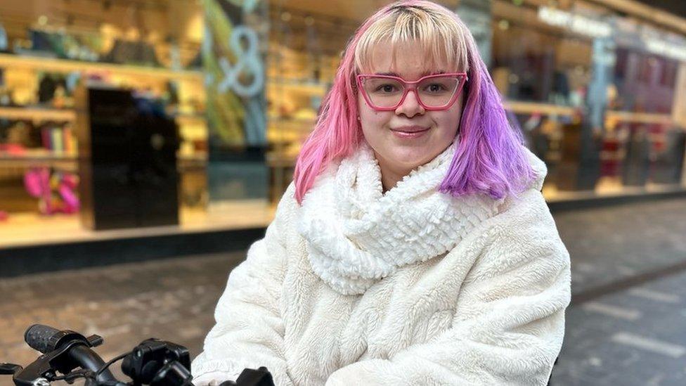 Sophia Dunn. Sophia is a 19-year-old white woman. She has hair dyed pink on one side and purple on the other with a blonde fringe. She has pink rimmed glasses framing her brown eyes and is smiling at the camera. She wears a fluffy cream jumper with matching scarf and is on a mobility scooter she uses. Sophia is pictured outside a clothing shop in Liverpool