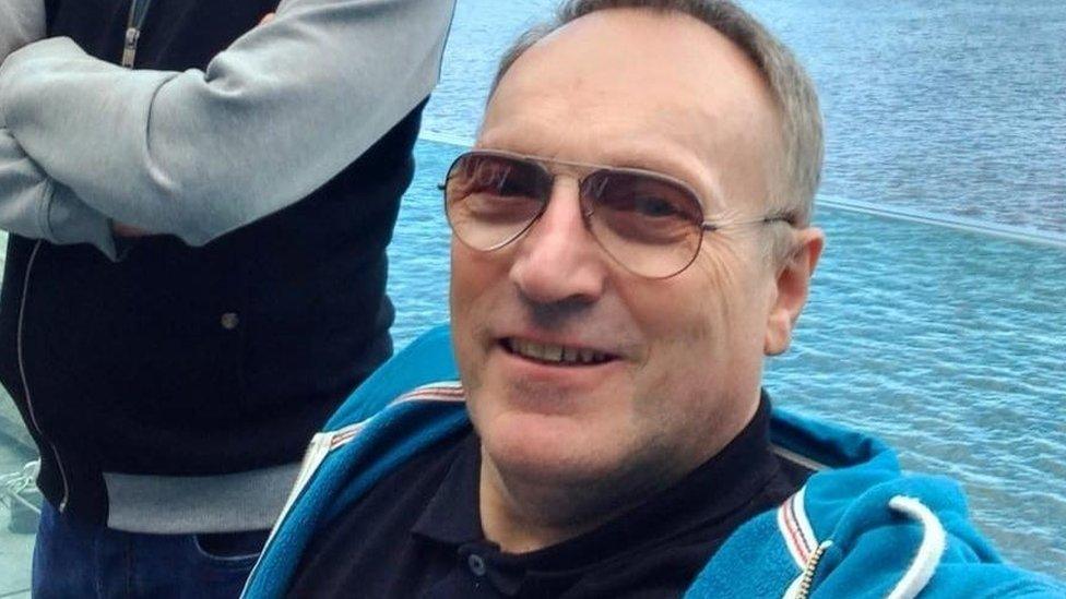 Man with glasses and short hair wearing a blue fleece sits with sea or a river behind him