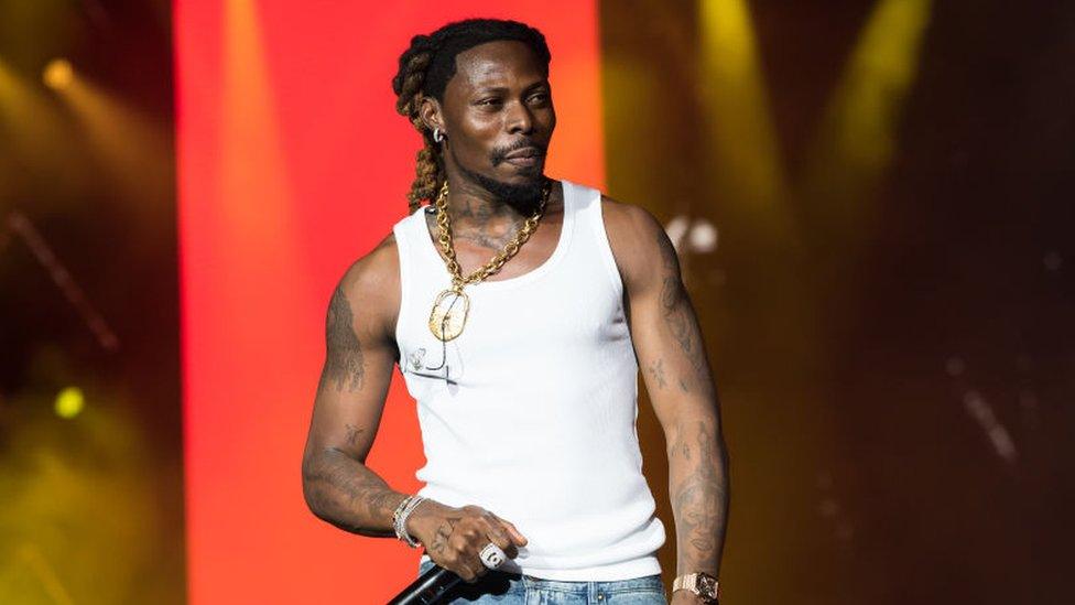 Asake performs onstage during day one of Afro Nation Miami in May. Asake, a black man in his 20s with braided hair tied back wears a white vest and a gold chain, holding a microphone in his right hand. He has a large diamond ring and watch and has heavily tattooed arms and neck. He is lit by stage lights and the staging behind him includes a bright red panel and yellow spot lights.