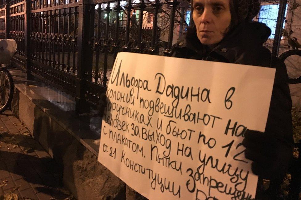 Woman holding sign saying "They hang Dadin by his handcuffs and 12 men beat him for taking to the streets with a protest banner. Torture is banned under Article 21 of the Russian constitution