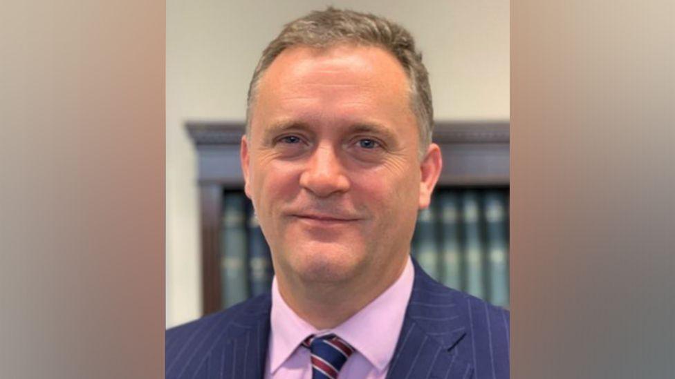 Lincoln Brookes, who has short grey hair and is smiling at the camera. He is wearing a navy, pinstriped suit jacket with a pink shirt and blue and red tie.