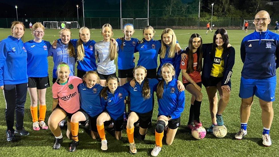 A group of girls poses for a photograph with woman and a man on a sports pitch In Newry