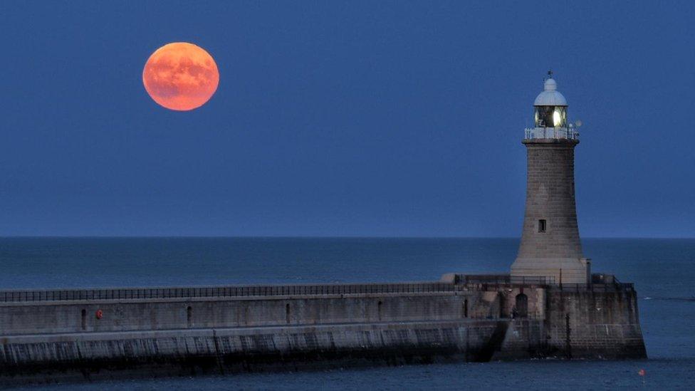 A bright full Moon, in the clear sky, above the sea, with a lighthouse in the foreground