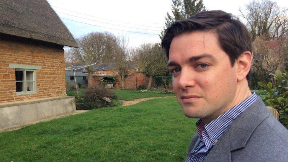 Duncan Greenaway with short dark hair and grey jacket sitting the garden of a brick-built cottage