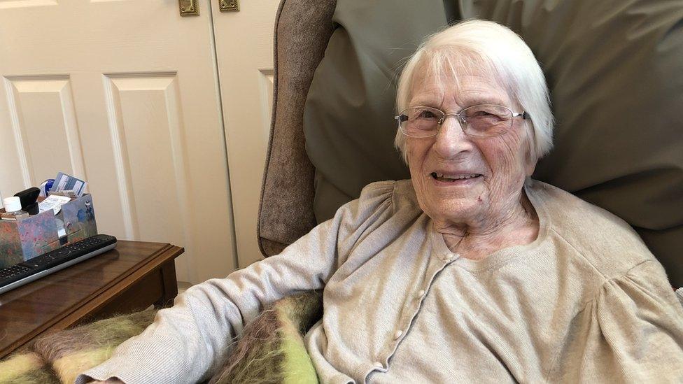 An older white woman with grey hair and glasses sitting in an armchair smiling