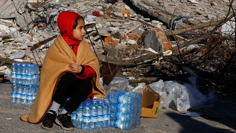 Child seated amid crates of water in the aftermath of earthquake in Kahramanmaras