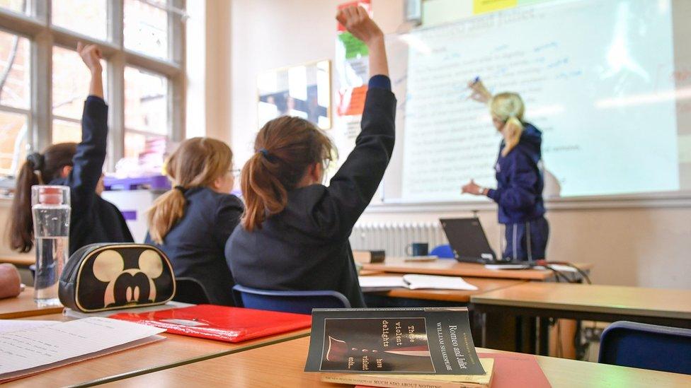 A school classroom with children raising their hands as a teacher writes on a board