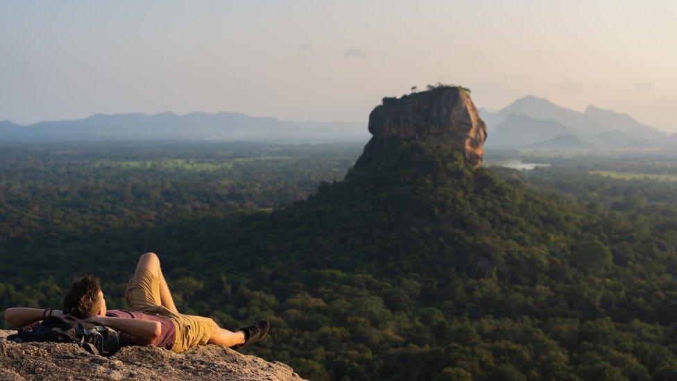 A tourist lying down, looking at Lion Rock across a valley of trees