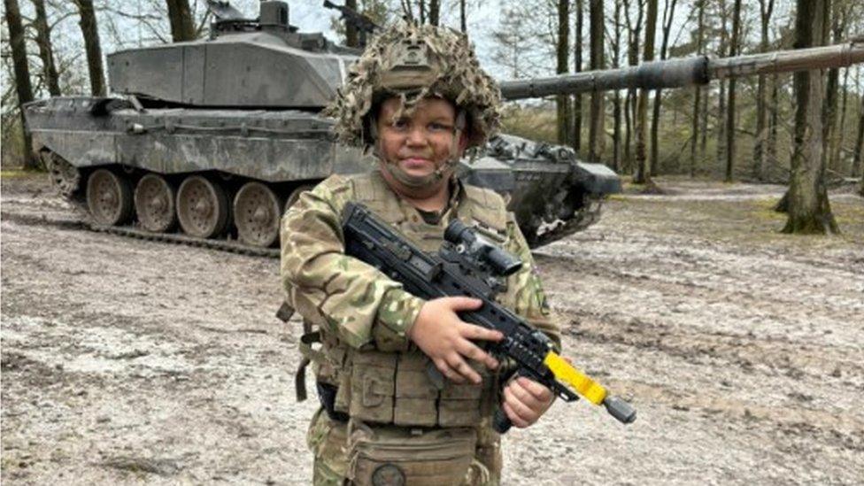 Noah kitted out in a soldier's uniform and carrying a rifle, standing in front of a tank