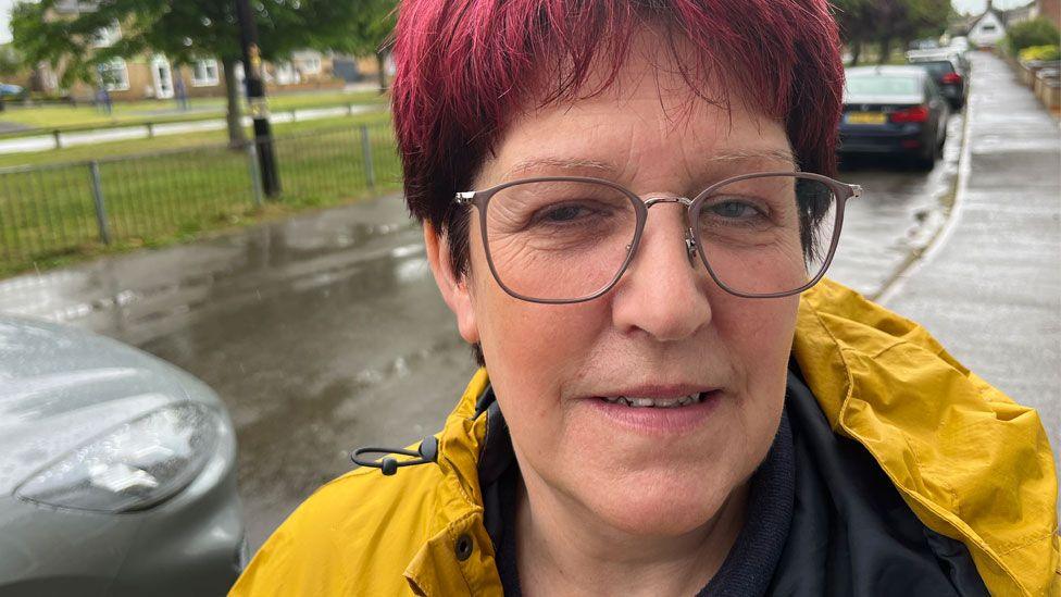 Sharon Stevens who has short bright pink hair and is wearing glasses. She has on a bright yellow rain jacket over a dark top. Behind her is a street with parked cars and on her right are railings and a park.