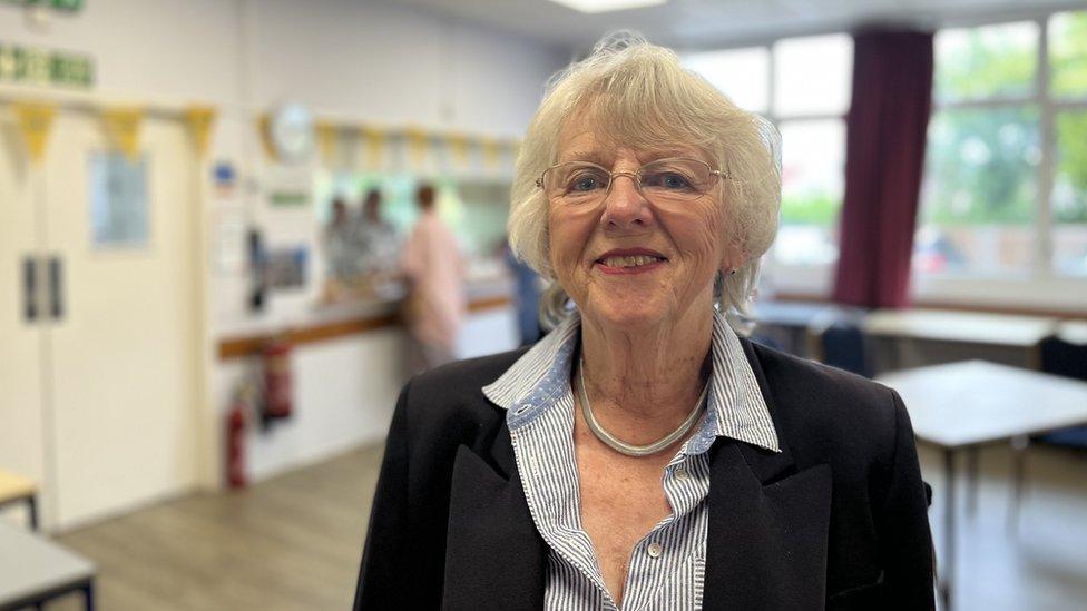 a white woman with grey hair wearing a dark blazer in a community hall