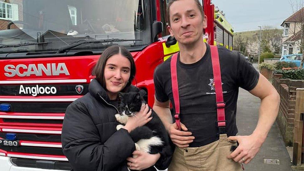 A woman holding a black and white cat with a firefighter