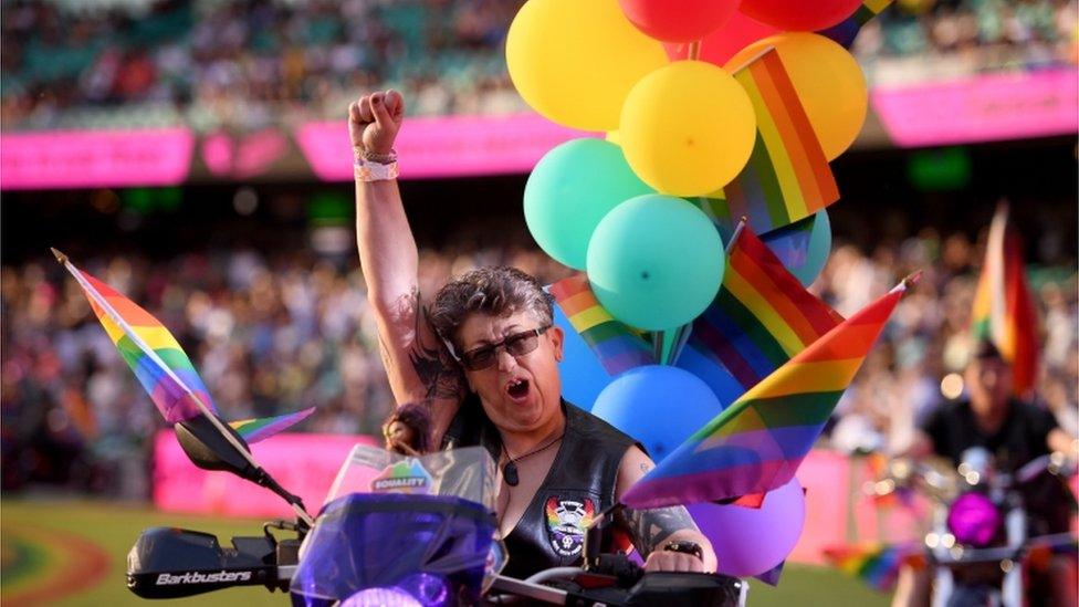 A female biker, with her fist in air, rides with rainbow flags and balloons in front of the crowd