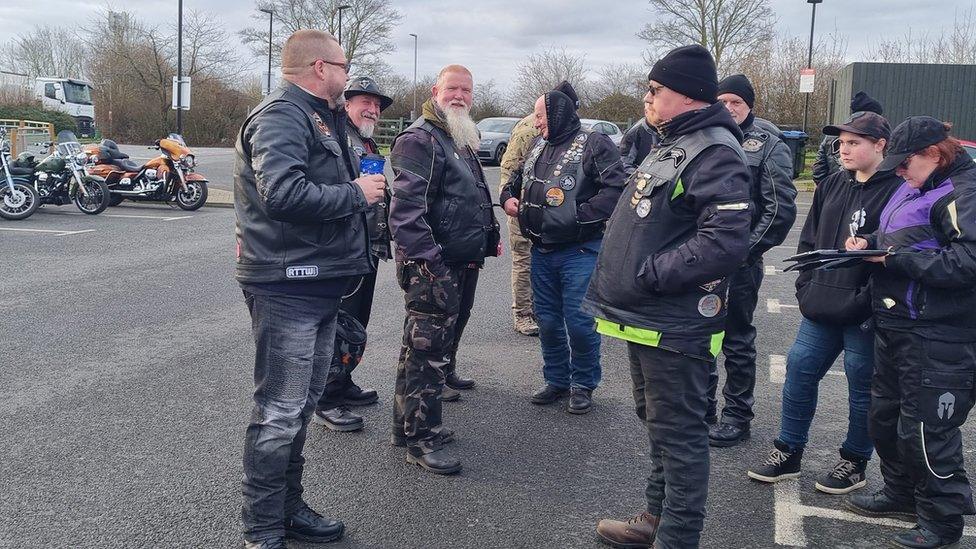 Bikers, some in leathers, standing in a car park with bikes in the background