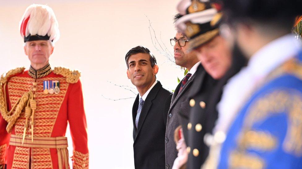 The UK Prime Minister smiles as he lines up alongside men in various uniforms.