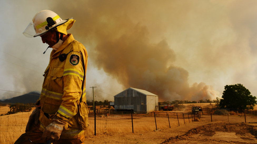 A lone firefighter stands with his head bowed against plumes of smoke in the distance from a bushfire