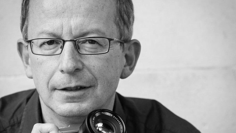 Ken Abbot smiling at the camera holding a camera in a black and white image