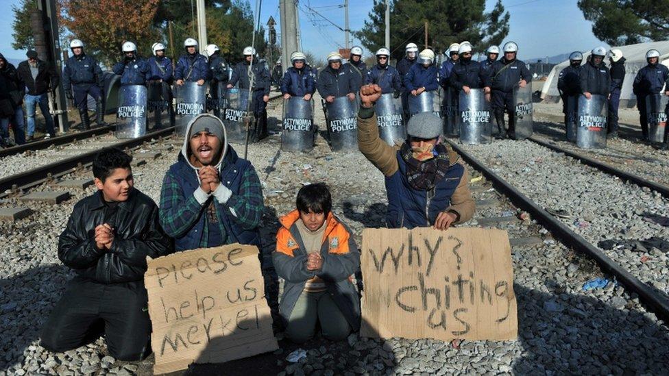 Greek police officers stand guard behind two men and two children demonstrating between railway tracks as they wait with other migrants and refugees to cross the Greek-Macedonian border near Idomeni on 7 December