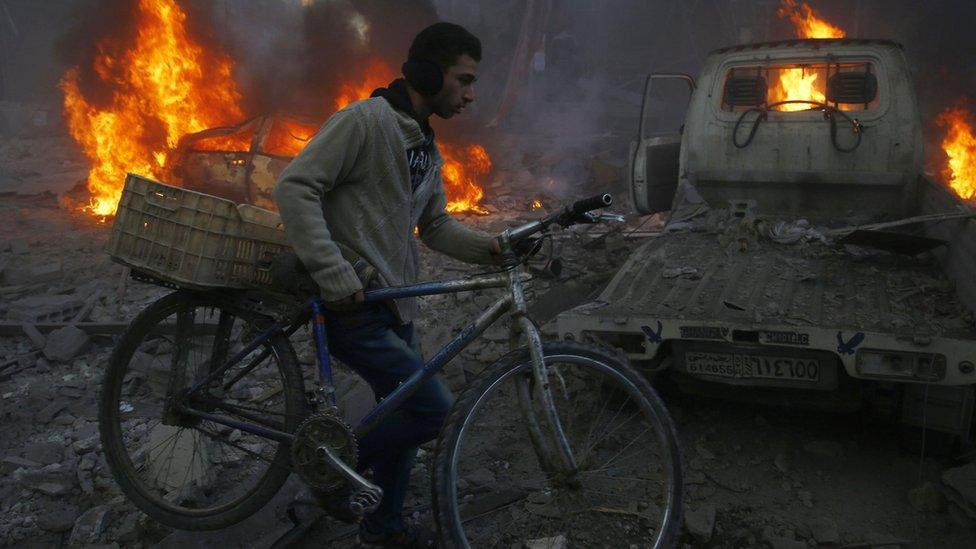 A man carries his bicycle past debris and burning cars following air strikes in the Damascus suburb of Hamouria (9 December 2015)