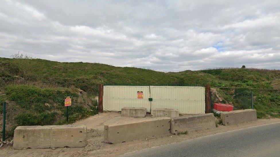 Roadside image of the site entrance, with concrete blocks and metal fencing.