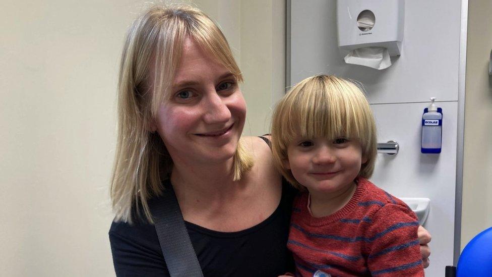 Laura and Sasho in a doctor's evaluation room. A sink with paper towels and soap can be seen behind them. Laura is blonde and wearing a black top. Her arm is around Sasho. Sasho is also blonde sat on Laura's knee, wearing a red and blue stripe jumper. Both of them are looking directly at the camera and smiling.