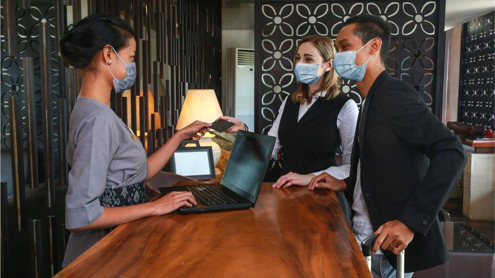 Couple and receptionist at counter in hotel wearing medical masks as precaution against virus. Young couple on a business trip doing check-in at the hotel