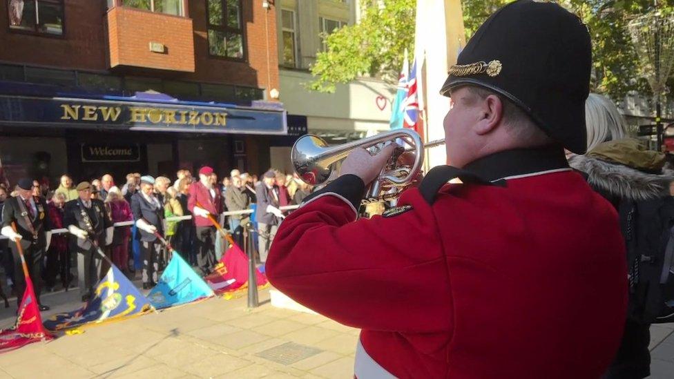 Man in red uniform with black hat plays the flugelhorn. Regimental standards are visible in the background