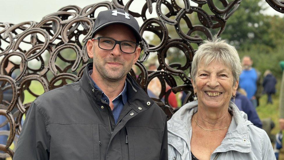 A man and a woman stand in front of a horse made of horseshoes