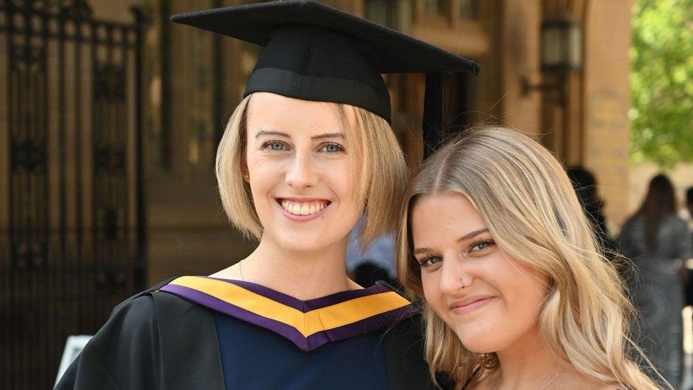 Laura and Gracie Nuttall at Laura's university graduation in the summer of 2022. Laura is on the left and is smiling. She has short blonde hair and she's wearing a navy and black university graduation gown and has a black mortarboard hat on. Gracie is to her right and she has longer, curly blonde hair and has a ringed nose piercing