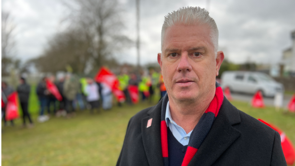 Mike Hobbs from Unite the union is looking straight at the camera and is wearing a black jacket and scarf