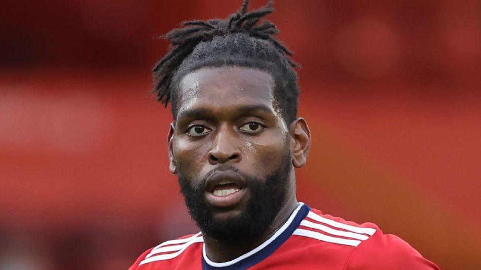 Close-up of Jay Emmanuel-Thomas in a red Aberdeen Football Club shirt. He has a dark beard and close-cropped moustache. His dreadlocks are tied up high in a ponytail on his head. 
