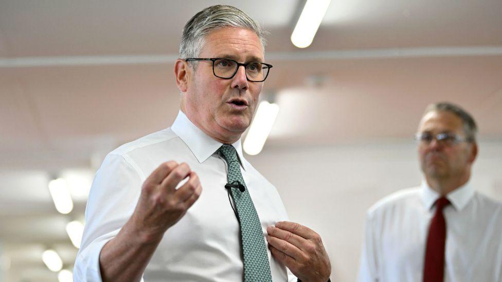 Keir Starmer, with grey hair and glasses, speaks with his right hand extended slightly in front of his body. He is wearing a white shirt and green tie.