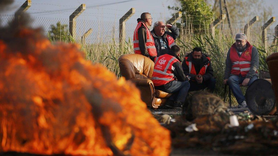 big flames in foreground as workers wearing CGT high-vis jackets sit in the background