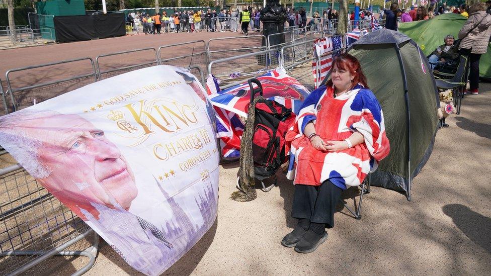 A woman in a Union Jack flag cape sits in a deck chair next to a flag of King Charles