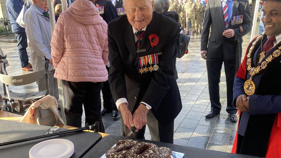 Ron Collins wearing a suit with poppy and medals stands next to Salisbury's mayor and cuts his birthday cake, which is shaped as the number 102