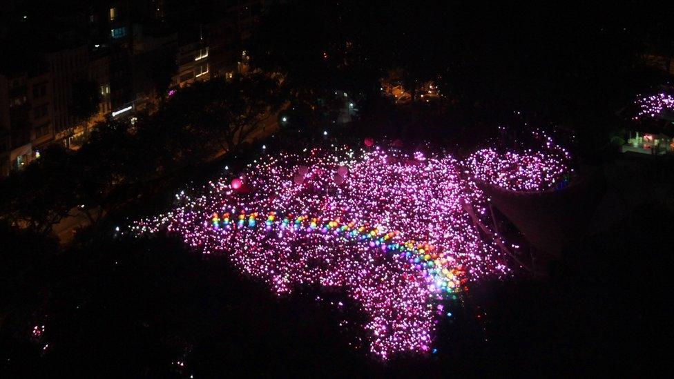 An aerial photo of the demonstration space in Singapore, filled with hundreds of pink lights in an irregular shape. Arcing across the centre is a series of coloured lights creating the image of a rainbow, a well-known LGBT symbol.