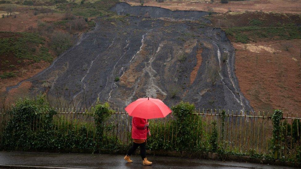 Dynes gydag ymbarel yn cerdded heibio tirlithriad tomen lo ym Mhendyrus, Rhondda Cynon Taf