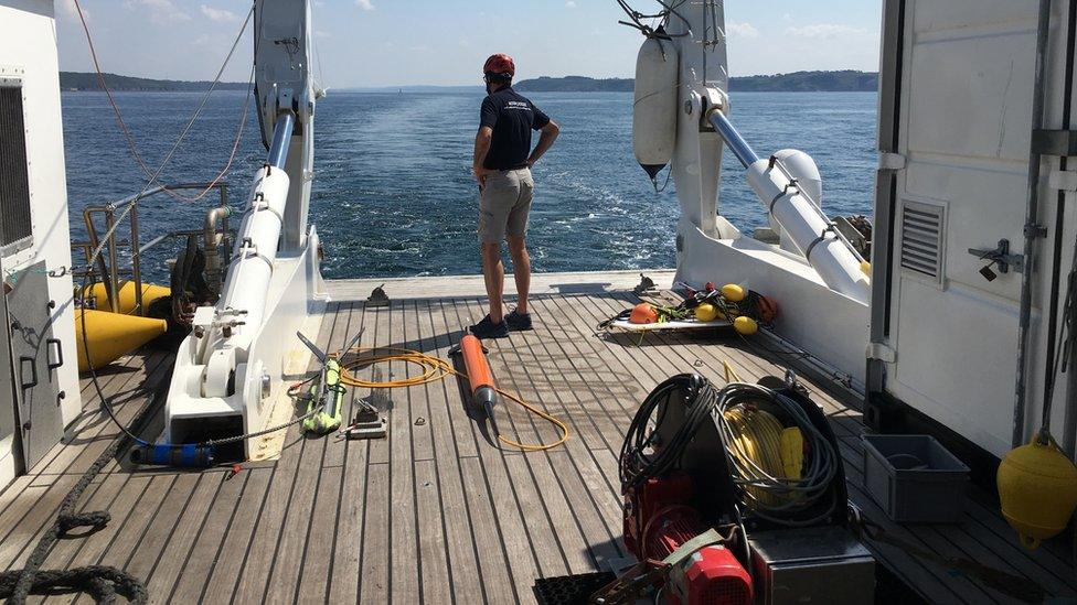 A helmeted crew member stands astern, looking at the waves, with piles of cables and other equipment pooled nearby