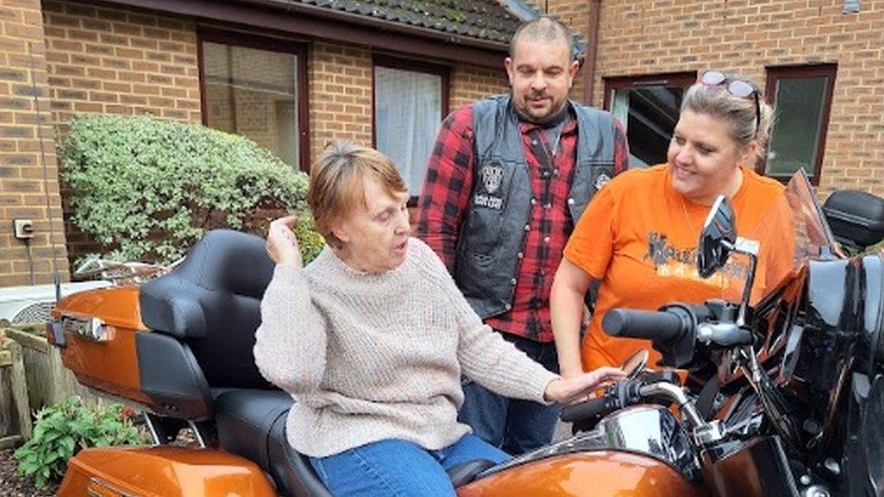 Older women with short red/brown hair sits on the seat of (possibly) a Harley Davidson, watched by two bikers