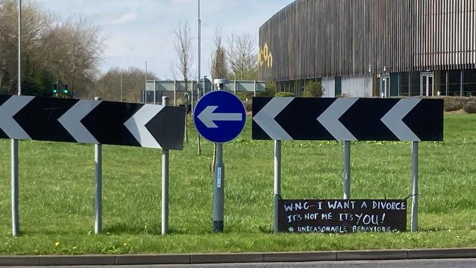 Black sign with white lettering on a roundabout. The sign says: "WNC... I want a divorce"