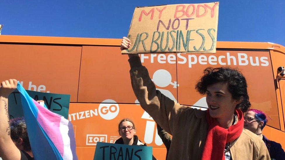 protestors surround the bus in Cambridge, one holds a transgender flag and another a sign that says 'My body not your business'