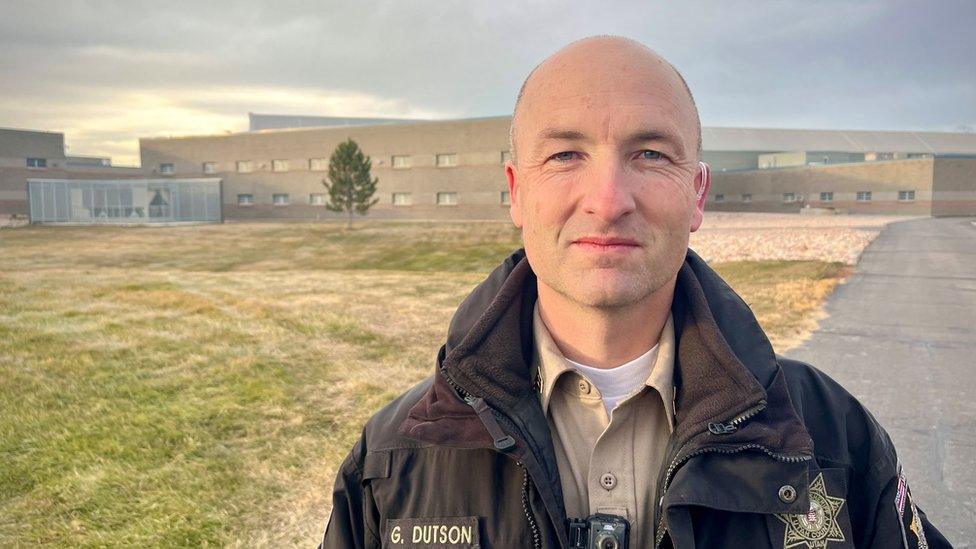 Man in a police uniform looking into the camera in the foreground with Utah County Jail in the background - a large sand coloured building