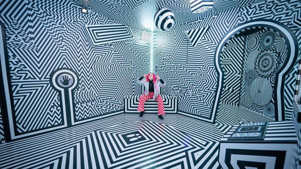 Woman with pink hair sitting inside room with jagged black and white stripes painted on the wall, ceiling and floor