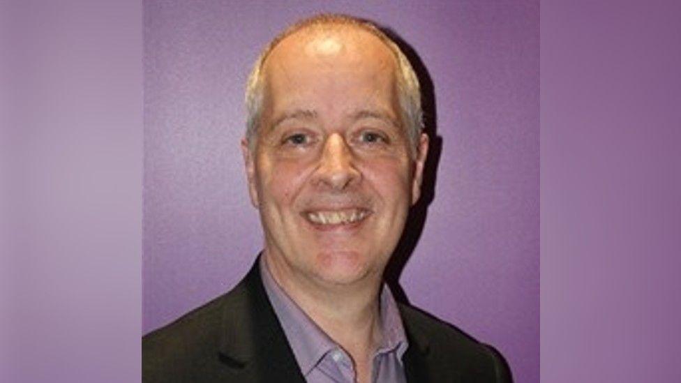 A head and shoulders photo of a bald man wearing a black blazer and a lilac shirt, smiling against a purple background.