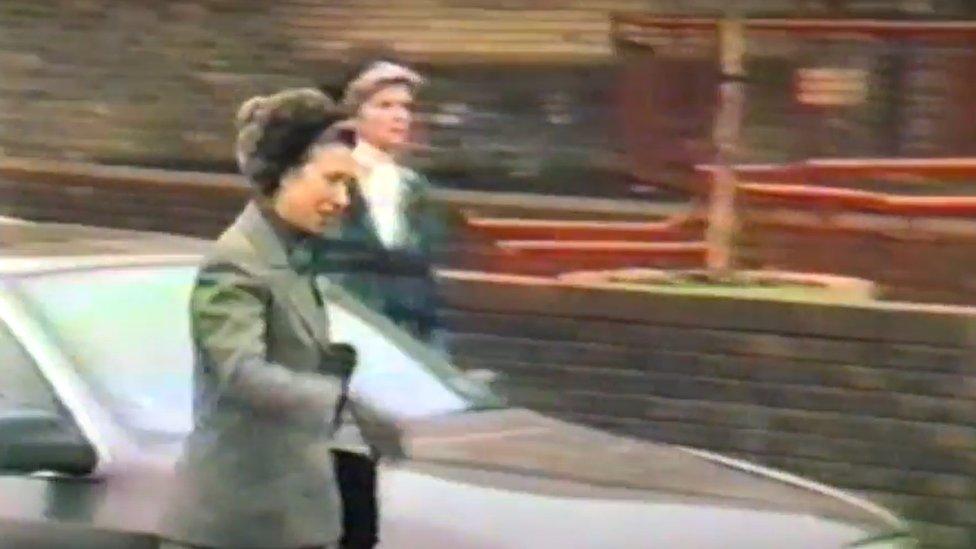 Woman wearing long grey coat and grey hat walks past a car outside a brick building with red railings