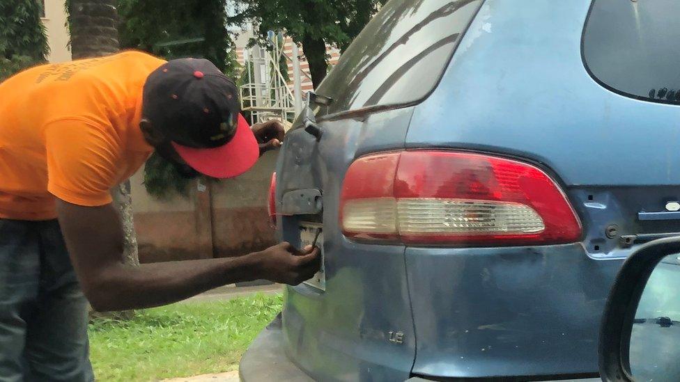 A man in an orange shirt removes a vehicle's number plate